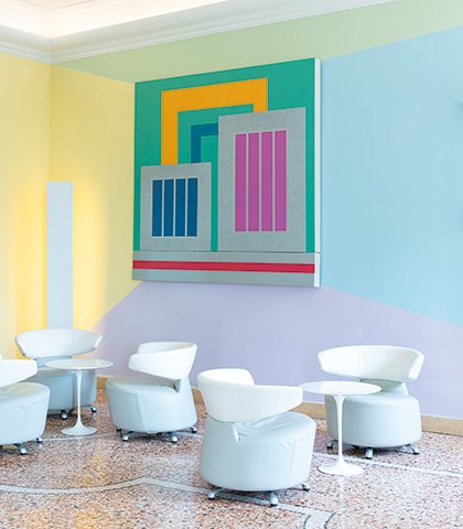Angolo lounge luminoso con poltrone bianche e pareti dai toni pastello decorate da un’opera geometrica di design