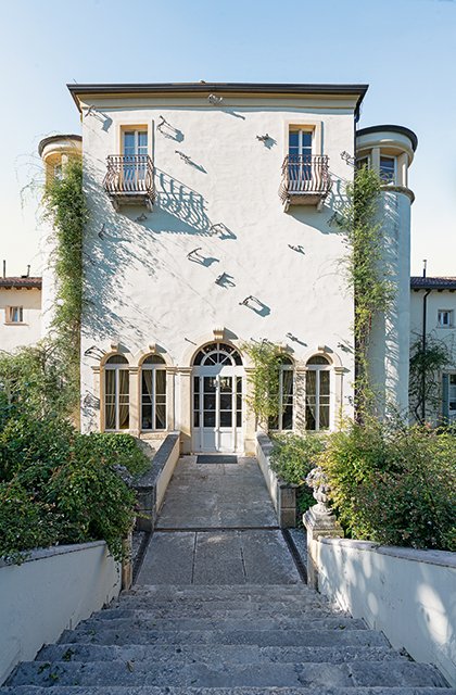 Facciata posteriore della villa con pareti chiare, rampicanti verdi e scale in pietra che conducono all’ingresso decorato da archi