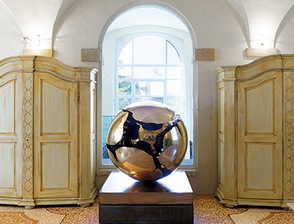 Scultura sferica dorata di Arnaldo Pomodoro esposta tra armadi antichi e luce naturale proveniente da una grande finestra