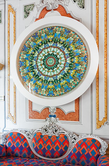 Opera circolare di Damien Hirst con motivi a farfalla in stile mandala, sopra divano barocco rivestito in tessuto geometrico colorato