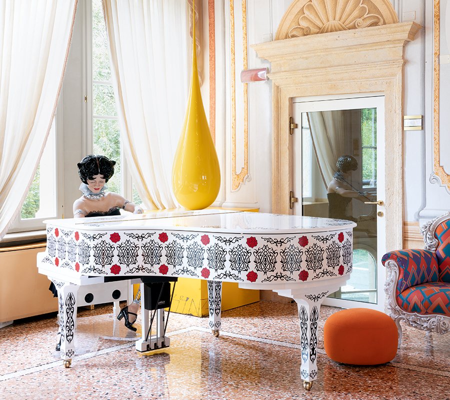 Dettaglio della hall del Byblos Art Hotel con pianoforte decorato e figura femminile artistica accanto a una scultura gialla a goccia.